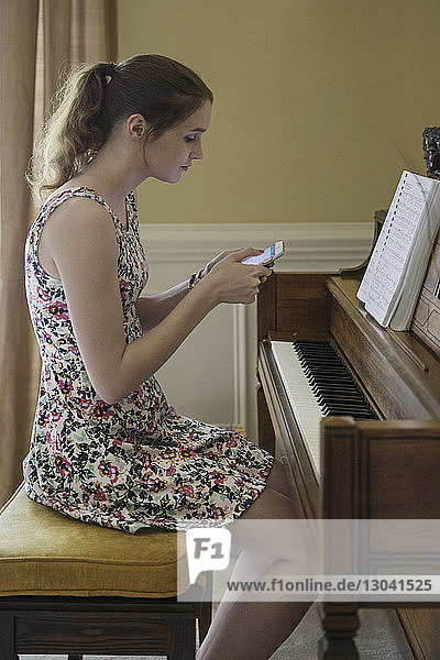 Seitenansicht einer Teenagerin  die ein Mobiltelefon benutzt  während sie zu Hause am Klavier am Tisch sitzt