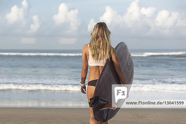 Rückansicht einer Frau  die ein Surfbrett trägt  während sie am Strand in Richtung Meer läuft
