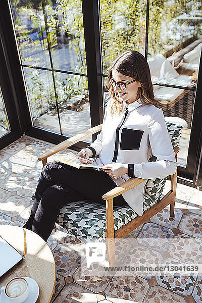 Schrägansicht einer Geschäftsfrau  die ein Mobiltelefon benutzt  während sie mit einem Buch im Café sitzt