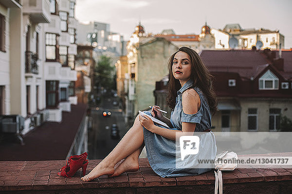 Porträt einer selbstbewussten jungen Frau mit Notizblock in Seitenansicht  die auf einer Stützmauer gegen Gebäude in der Stadt sitzt