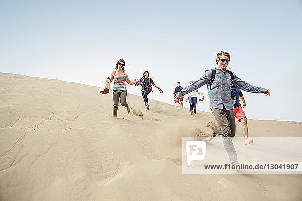 Cheerful friends running down sand dune against clear sky