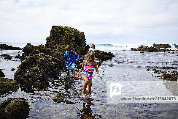 Brother and sister walking in water by rocks at Laguna Beach