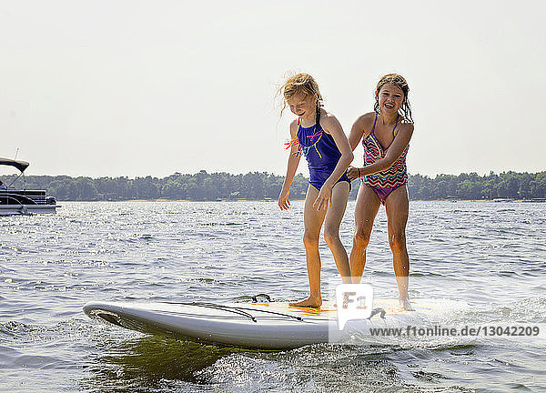 Cheerful siblings standing on paddleboard in lake against clear sky during sunny day