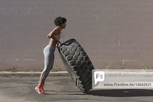 Full length of woman lifting tire truck while exercising against wall