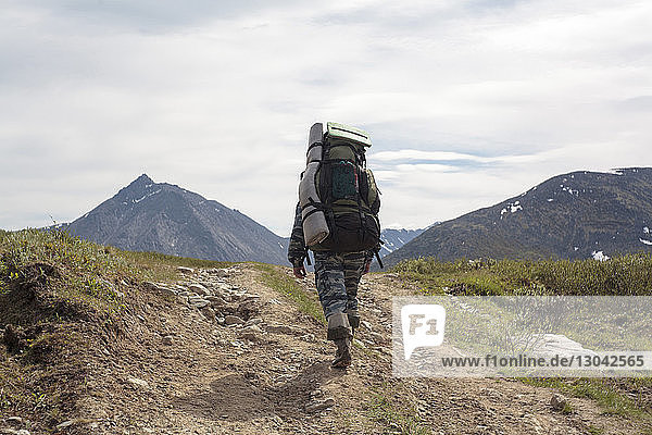 Rear view of male hiker with backpack walking on mountain against cloudy sky