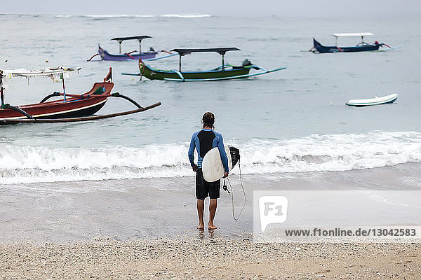 Rear view of man looking at sea while carrying surfboard on shore