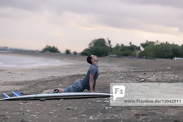 Side view of man performing cobra pose by surfboard at beach