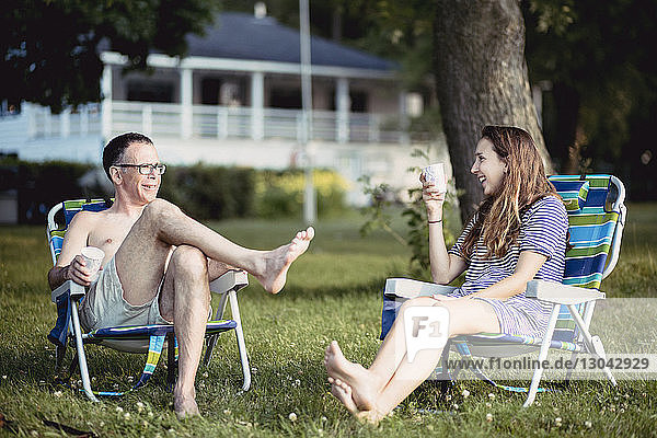 Father and daughter smiling while sitting on deck chairs in lawn