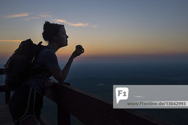 Weibliche Wanderin isst Apfel  während sie bei Sonnenuntergang am Beobachtungspunkt gegen den Himmel steht