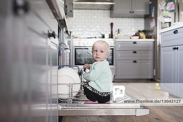 Cute girl looking away while sitting in dishwasher at kitchen