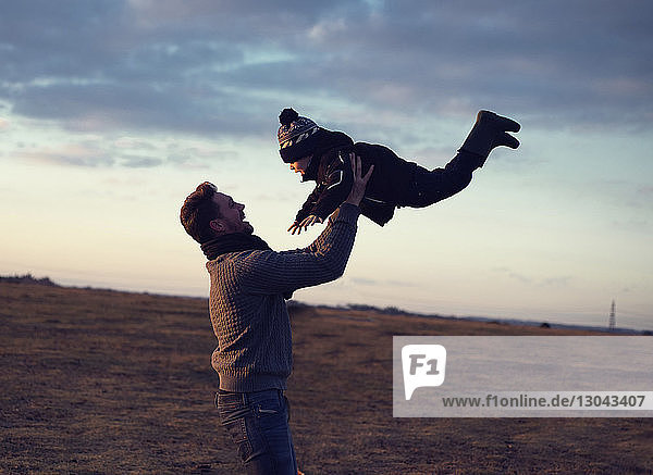 Vater spielt mit seinem Sohn  während er ihn auf dem Feld gegen den bewölkten Himmel hebt