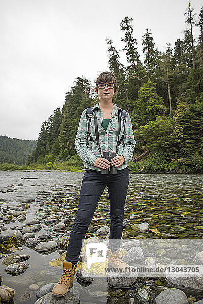 Portrait of female hiker holding binoculars while standing on rocks over stream at Redwood National and State Parks