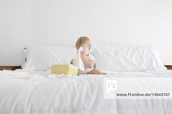 Junge versteckt Gesicht mit Gesichtsgewebe beim Spielen im Bett zu Hause