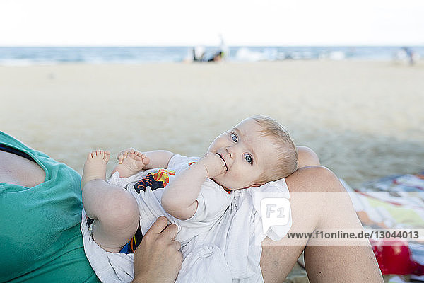 Porträt eines süßen Sohnes mit Fingern im Mund auf dem Schoß der Mutter am Strand liegend