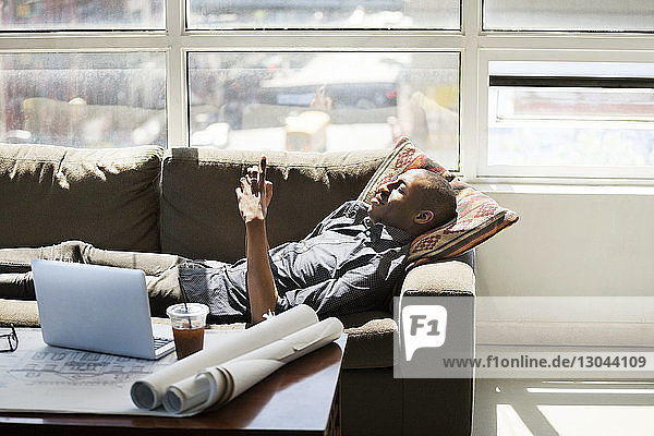 Businessman using smart phone while lying on sofa in creative office