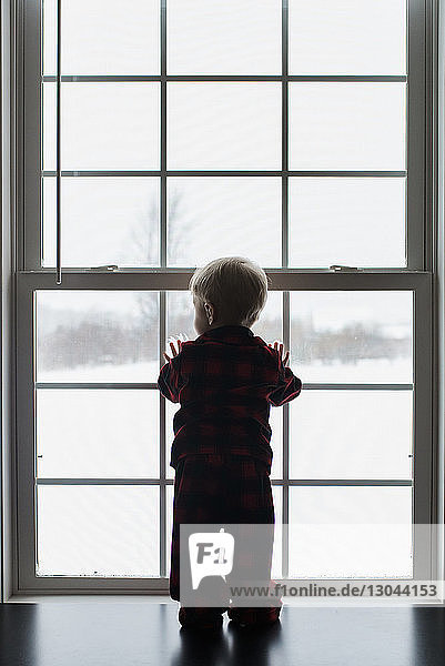 Rückansicht eines zu Hause stehenden Jungen  der durch ein Fenster schaut