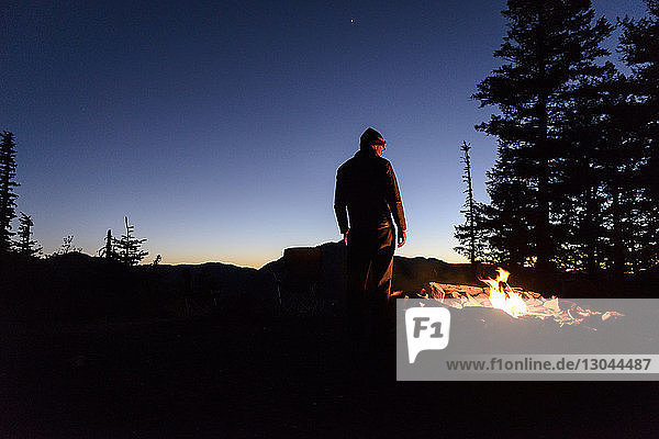 Rückansicht eines am Lagerfeuer stehenden Mannes vor blauem Nachthimmel