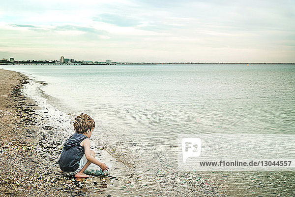Junge hält Schüssel mit Kieselsteinen in der Hand  während er am Strand kauert
