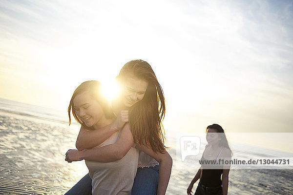 Teenagermädchen mit Huckepack-Freundin bei Sonnenuntergang am Strand