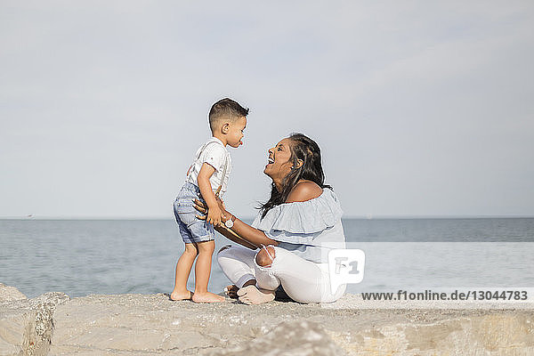 Seitenansicht eines glücklichen Sohnes und einer glücklichen Mutter  die spielen  während sie auf einer Stützmauer gegen das Meer sitzen