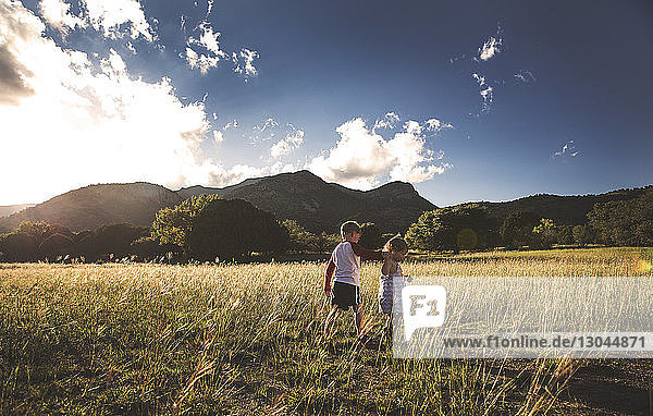 Siblings walking on grassy field by mountains against sky
