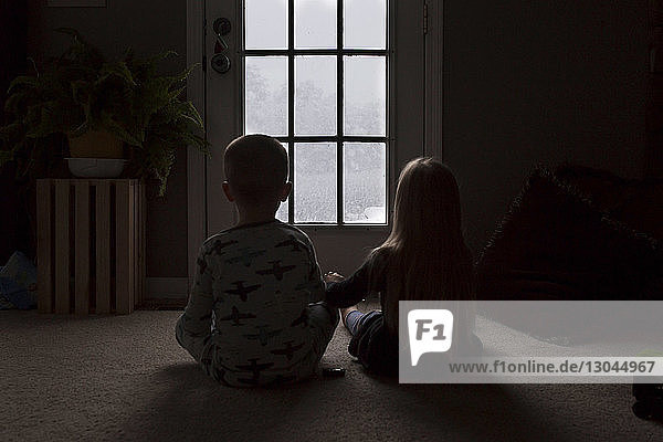 Rückansicht von Geschwistern  die durch das Fenster schauen  während sie in der Dunkelkammer auf dem Boden sitzen