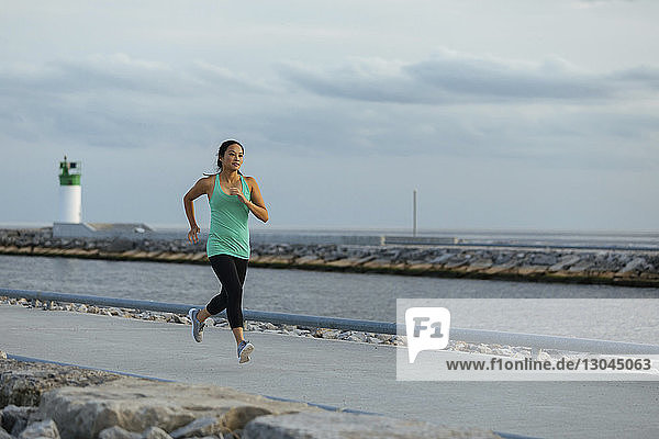 Eine Frau in voller Länge joggt auf einem Fußweg am Meer gegen den Himmel