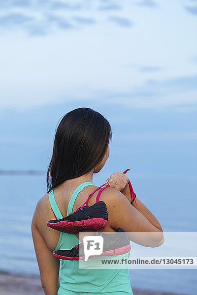Rückansicht einer nachdenklichen Frau  die Sportschuhe hält  während sie am Strand steht