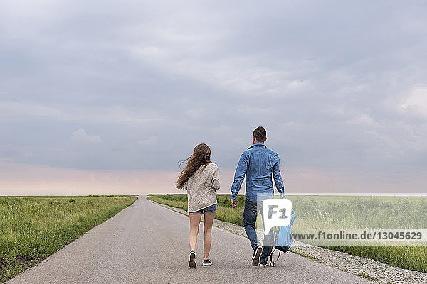 Full length rear view of hiking couple walking on country road amidst grassy field against sky