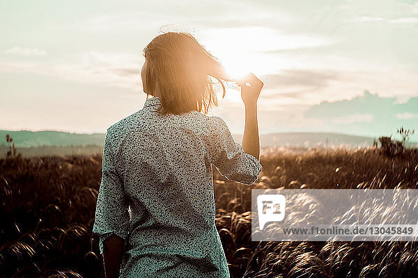 Rückansicht einer Frau  die Haare hält  während sie bei Sonnenuntergang auf dem Feld gegen den Himmel steht