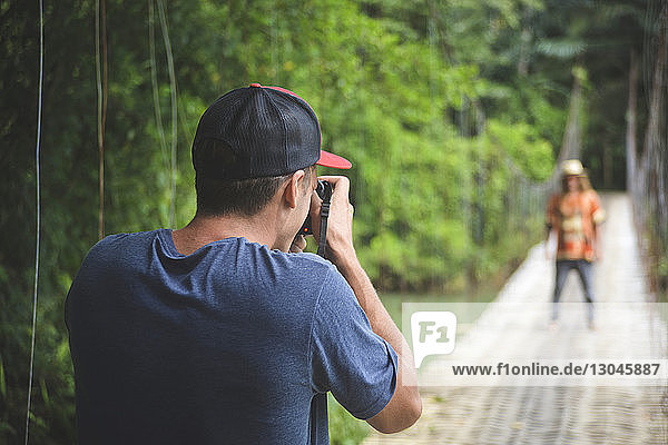 Rear view of man photographing friend while standing on rope bridge