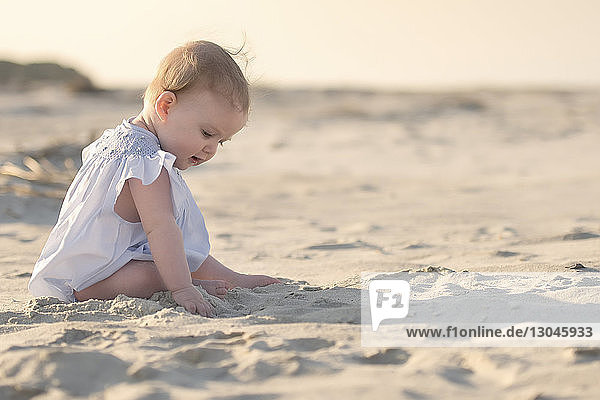 Side view of baby girl sitting on sand at beach against sky during sunset