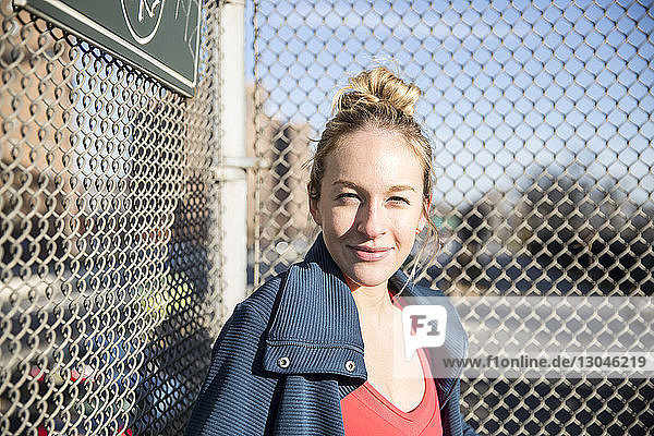 Portrait of female athlete standing by fence in city during sunny day