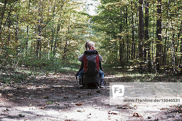Rear view of girl with backpack sitting on bag at dirt road amidst forest