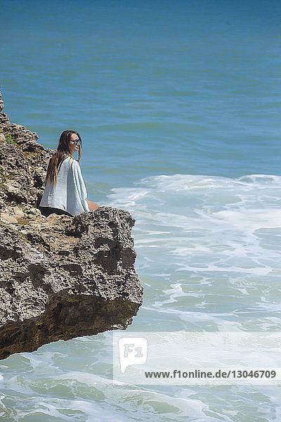 Hochwinkelaufnahme einer Frau  die an einem sonnigen Tag auf einem Felsen über dem Meer sitzt