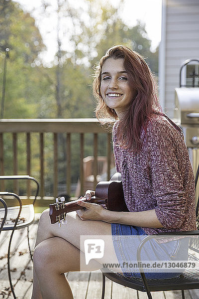 Porträt einer glücklichen Teenagerin  die Gitarre spielt  während sie auf einem Stuhl in der Veranda sitzt