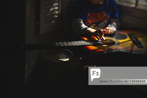 Low section of boy playing guitar while sitting in darkroom at home