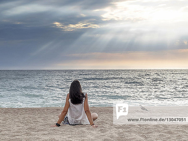 Rückansicht einer Frau  die auf das Meer schaut  während sie sich im Sonnenuntergang auf Sand vor bewölktem Himmel entspannt