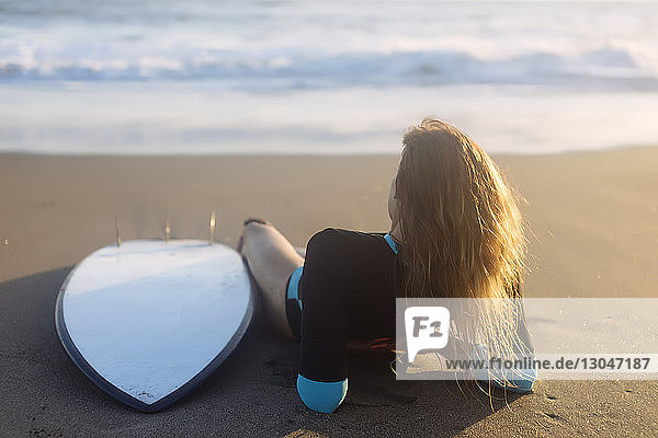Rückansicht einer Frau  die sich bei Sonnenuntergang am Strand am Surfbrett entspannt