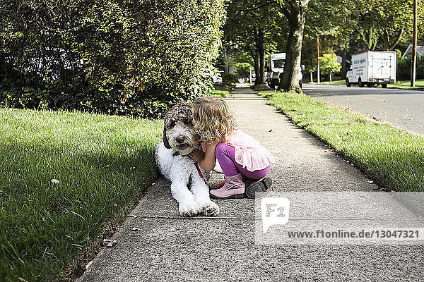 Seitenansicht eines Mädchens  das einen Hund küsst  während es auf einem Fußweg an der Straße kauert