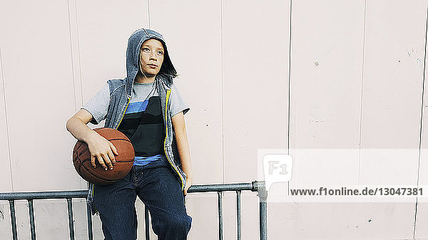 Junge mit Kapuzenhemd hält Basketball  während er auf einem Geländer an der Wand sitzt
