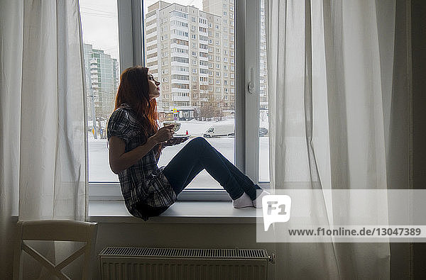 Seitenansicht eines nachdenklichen Teenagers  der eine Teetasse hält  während er zu Hause am Fenster sitzt