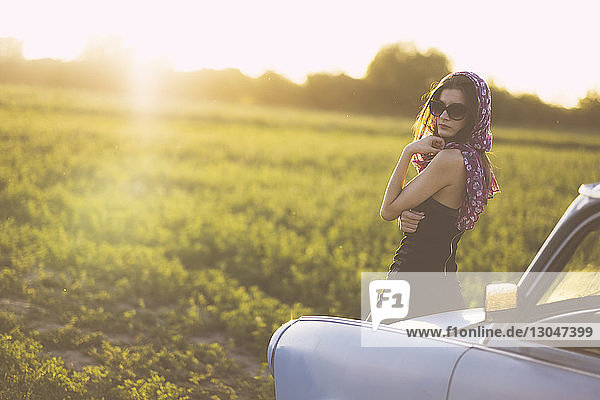 Porträt einer Frau mit Sonnenbrille  die bei Sonnenuntergang im Auto auf einem Grasfeld gegen den Himmel steht