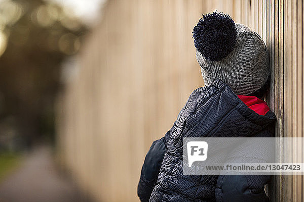 Seitenansicht eines Jungen in warmer Kleidung  der sich im Hinterhof an einen Holzzaun lehnt