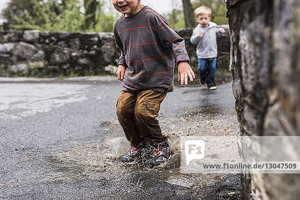 Midsection of boy jumping in puddle with brother in background