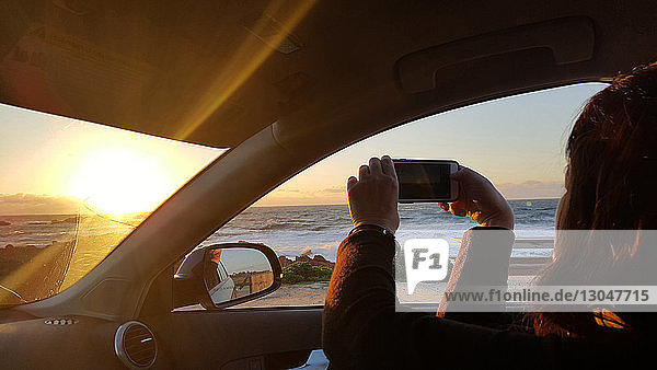 Boy photographing beach while sitting in car during sunset