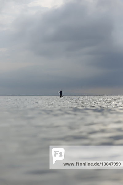 Blick aus mittlerer Entfernung auf den Silhouettenmann beim Paddeln im Meer vor bewölktem Himmel