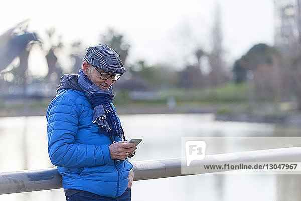 Man using mobile phone while standing by railing against lake at park