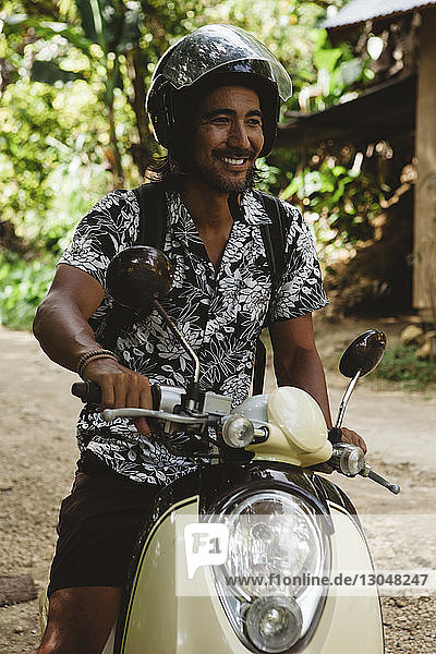 Lächelnder Mann mit Helm beim Fahren mit dem Motorroller auf unbefestigter Straße