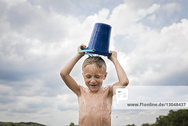 Shirtless boy pouring bucket of water over head against cloudy sky